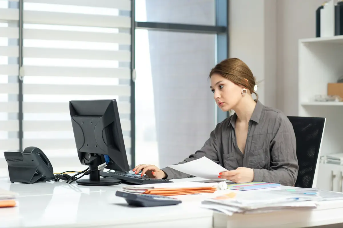 A woman working at an office computer with paperwork in hand and a desk phone beside her monitor