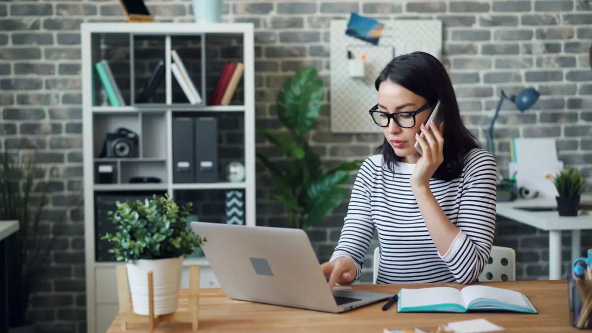 A woman wearing glasses taking a phone call while working at her laptop in a bright office